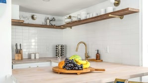 Una cucina con piastrelle bianche, mensole in legno, una ciotola di frutta con uva, banane e un melone, e una macchina per il caffè.