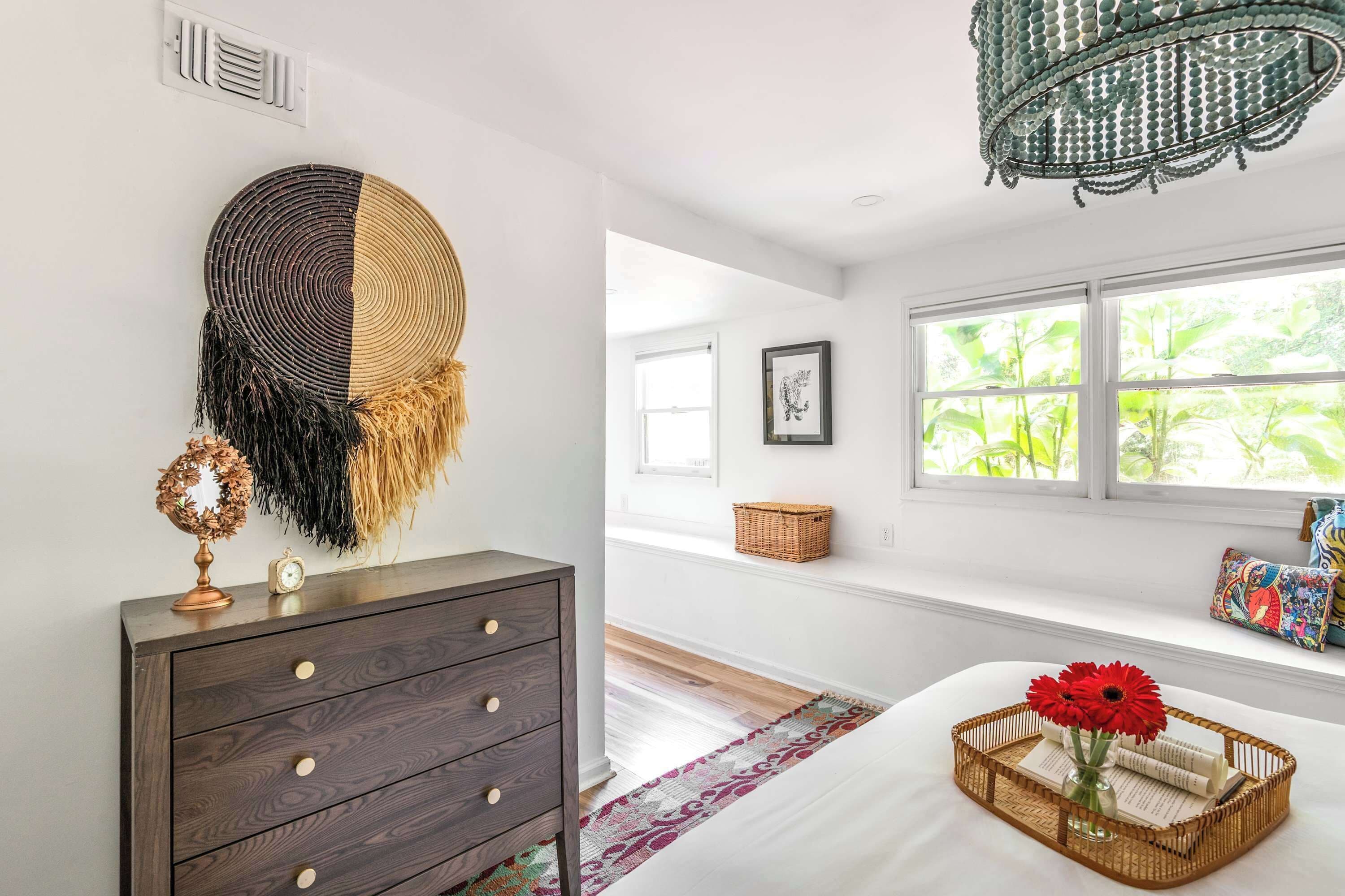 A bedroom with a wooden dresser, a woven wall hanging, a chandelier, a window with a view of greenery, and a bedside table with a basket and flowers.