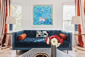 A blue sofa with a geometric patterned rug, a framed artwork above, and a vase with flowers on a side table.