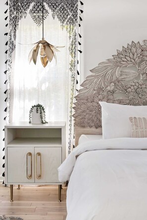 A bedroom with a white bed, a decorative headboard, a hanging light fixture, and a white nightstand with a plant. - Brooklyn Bungalow (Atlanta)
