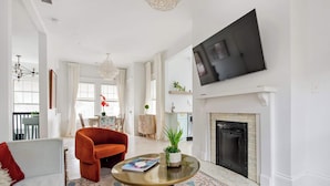 A spacious living room with a fireplace, a large flat-screen TV mounted above it, and a comfortable seating area with a sofa and an orange armchair. - Lace and Leather (Atlanta)