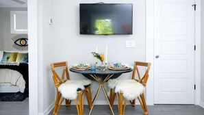 Uma sala de jantar com uma mesa redonda, duas cadeiras, uma televisão fixada na parede e uma porta branca.