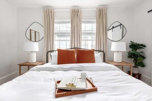 A neatly made bed with white linens, a tray with a book and a cup, and two bedside tables with lamps and a plant.