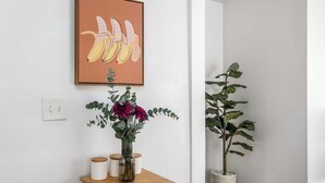 Una habitación con una mesita de madera, un florero con flores, una planta en maceta y un cuadro en la pared.