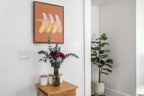 A room with a wooden side table, a vase with flowers, a potted plant, and a framed picture on the wall. - A Hard Day's Night (Atlanta)