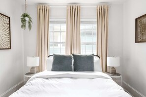 A neatly made bed with a white comforter and two gray pillows, flanked by two bedside tables with lamps, in a room with beige curtains and a hanging plant. - A Hard Day's Night (Atlanta)