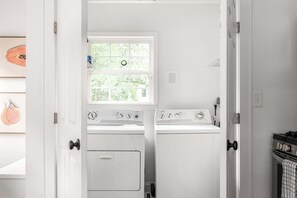 A laundry room with two white washers and dryers, a window with a view of greenery, and a door leading to another room. - A Hard Day's Night (Atlanta)