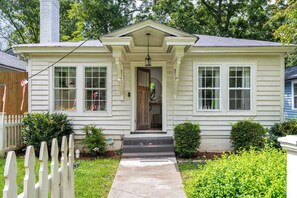 A single-story house with a front porch, a chimney, and a small front yard with a picket fence and bushes.