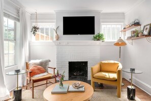 A living room with a white fireplace, a mounted TV, a yellow armchair, and a round coffee table with books and a vase.