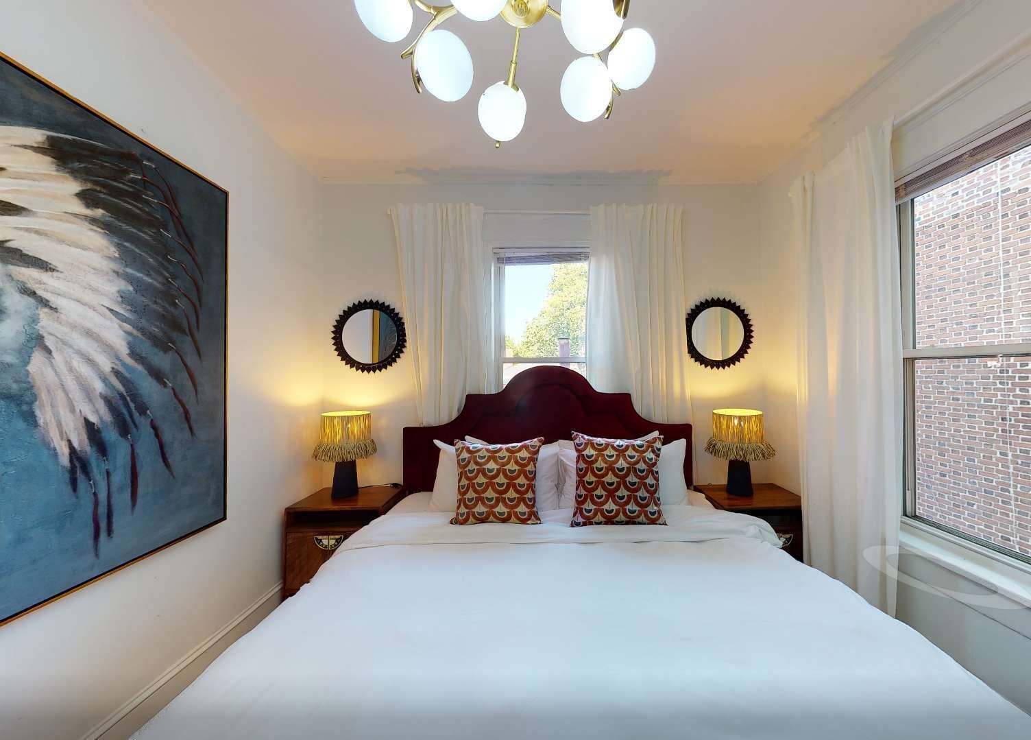 Un dormitorio con una cama grande, dos mesitas de noche con lámparas, un cuadro grande, dos espejos en la pared y una ventana con cortinas.