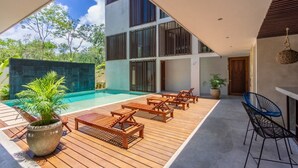 A modern outdoor pool area with wooden loungers and a concrete deck, surrounded by a multi-story building with large windows and a hanging light fixture. - New Luxe Spacious Loft ! Private Pool & Garden!! (Tulum)