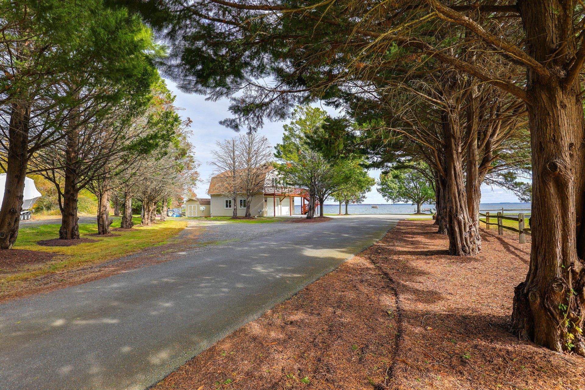A residential area with a paved driveway leading to a house with a red roof, surrounded by trees and a body of water in the background.