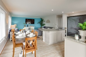 Una cocina moderna con una isla central, un comedor con mesa para cuatro y una sala de estar con sofá y televisor.