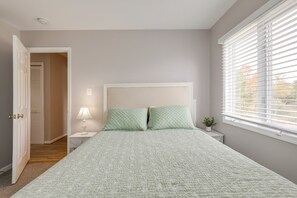 A bedroom with a bed, a nightstand, a lamp, a window with blinds, and a door leading to another room.