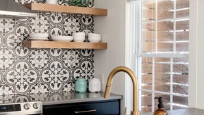A kitchen with a modern design, featuring a stainless steel sink, a black and gold faucet, and a patterned backsplash. There are wooden shelves above the sink holding dishes and a green vase. - Bohemian Bliss (Atlanta)