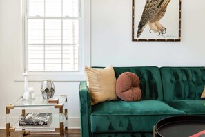 Una habitación con un sofá de terciopelo verde, una mesa de centro de vidrio y una ilustración enmarcada de un pájaro en la pared.