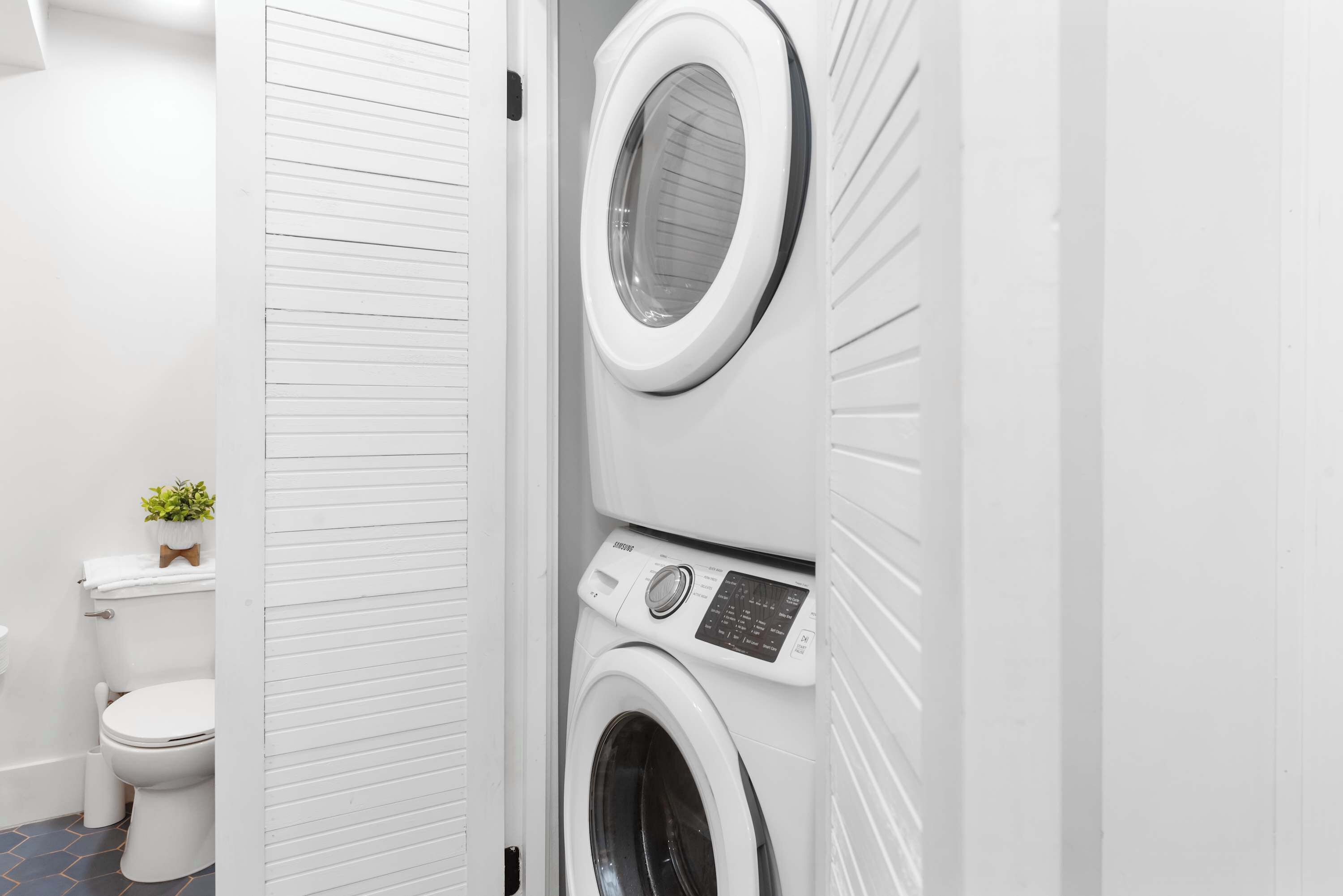 Une petite buanderie comprenant une machine à laver et un sèche-linge superposés, des toilettes et une petite plante en pot.