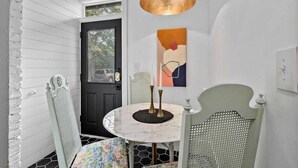 A dining area with a round table, two chairs, a black and white patterned table runner, a wall-mounted abstract painting, and a pendant light.