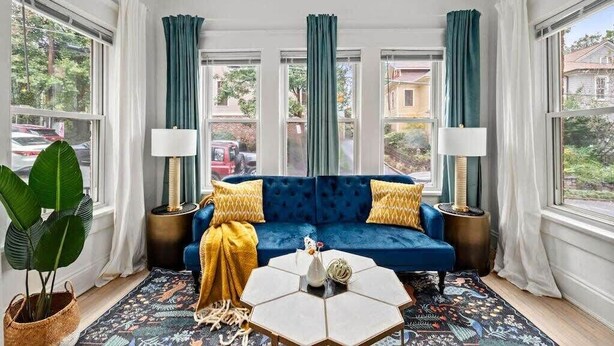 A room with a blue sofa, two lamps, a hexagonal coffee table, and a large window with curtains.