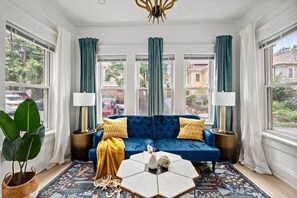 A room with a blue sofa, two lamps, a hexagonal coffee table, and a large window with curtains. - Field of Green (Atlanta)