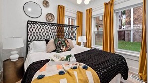 A bedroom with a black metal headboard, a bed with a white and yellow blanket, and a view of a brick building through large windows.