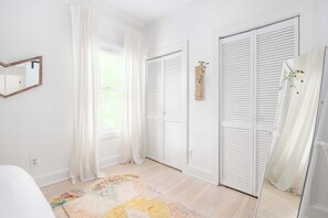 Quarto com cama, janela com cortinas, porta com venezianas e espelho na parede.