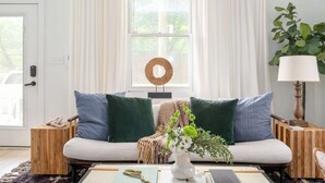 A living room with a sofa, two green pillows, a white vase with flowers, a wooden side table, and a lamp.