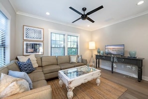 A living room with a sectional sofa, a glass coffee table, a ceiling fan, and a TV on a dark console.