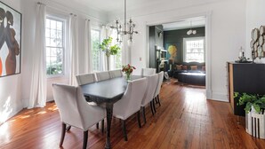 A dining area with a dark wooden table and white upholstered chairs, a chandelier, and a view into a living room with a sofa and a person in the background.