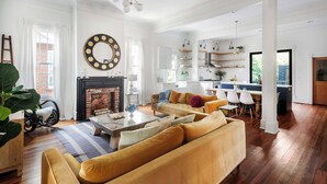 A spacious living room with a fireplace, a sofa, and a coffee table. There is a kitchen area with a dining table and chairs visible in the background.