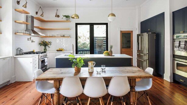 A modern kitchen with white cabinetry, wooden shelves, and a central island. There are hanging pendant lights, a built-in oven, and a refrigerator.