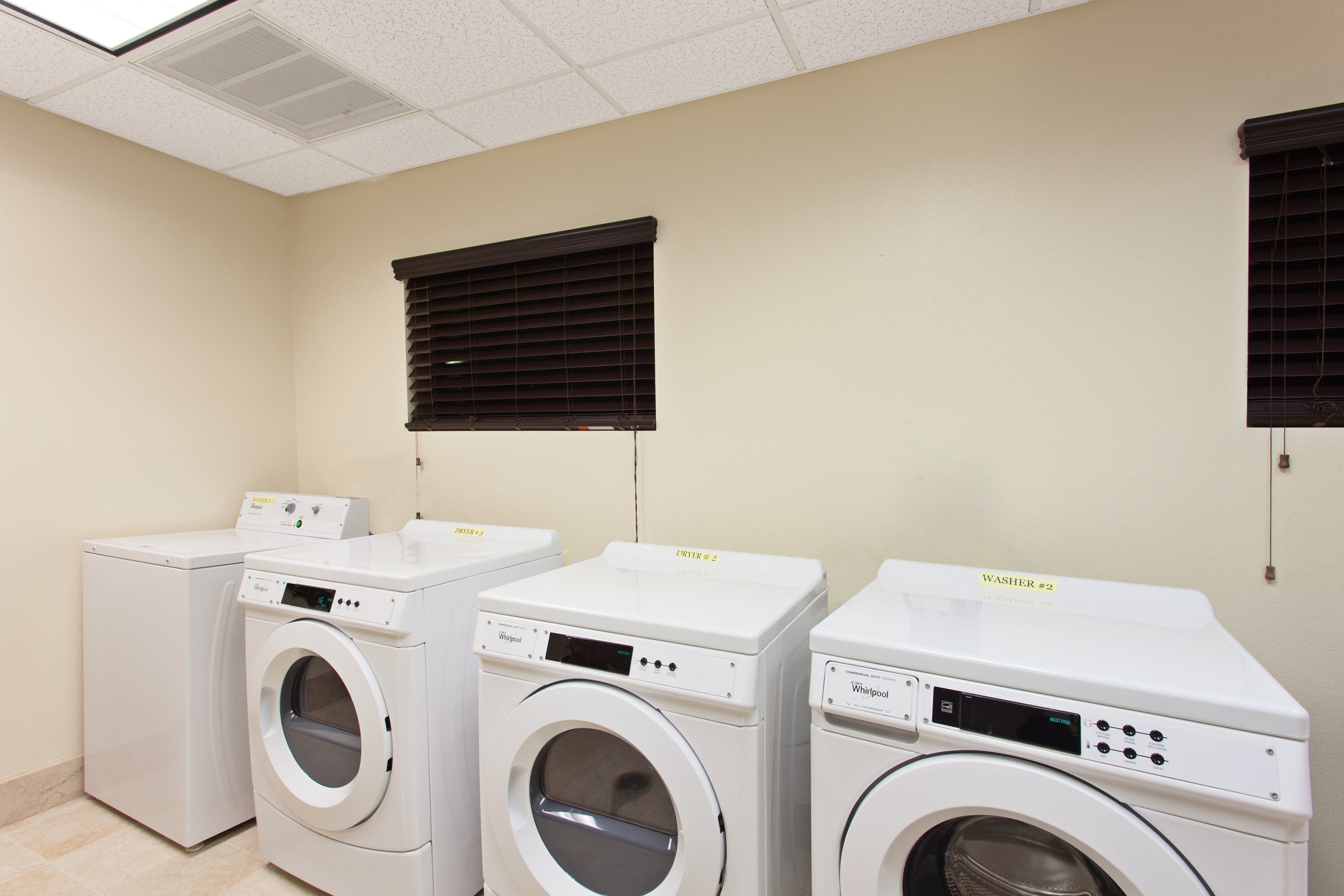 Une buanderie équipée de trois lave-linge et trois sèche-linge à chargement frontal, d’un plafond doté de luminaires fluorescents et de stores noirs aux fenêtres.