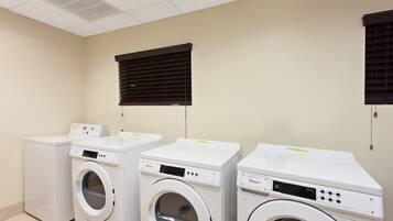 Une buanderie équipée de trois lave-linge et trois sèche-linge à chargement frontal, d’un plafond doté de luminaires fluorescents et de stores noirs aux fenêtres.
