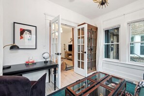 A modern living room with a glass coffee table, a black desk with a lamp, a framed artwork on the wall, and a view of the street through large windows. - Field of Gold (Atlanta)