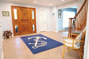 Un couloir avec une porte en bois, un tapis bleu orné d’une ancre blanche, une chaise en osier et un escalier.
