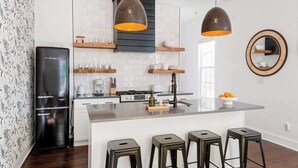 A modern kitchen with a black refrigerator, wooden shelves, a central island with stools, pendant lighting, and a mirror on the wall. - Whiskey Neat (Atlanta)
