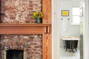 A brick fireplace with a wooden mantel, a vase with sunflowers, and a vintage bathtub in a bathroom.