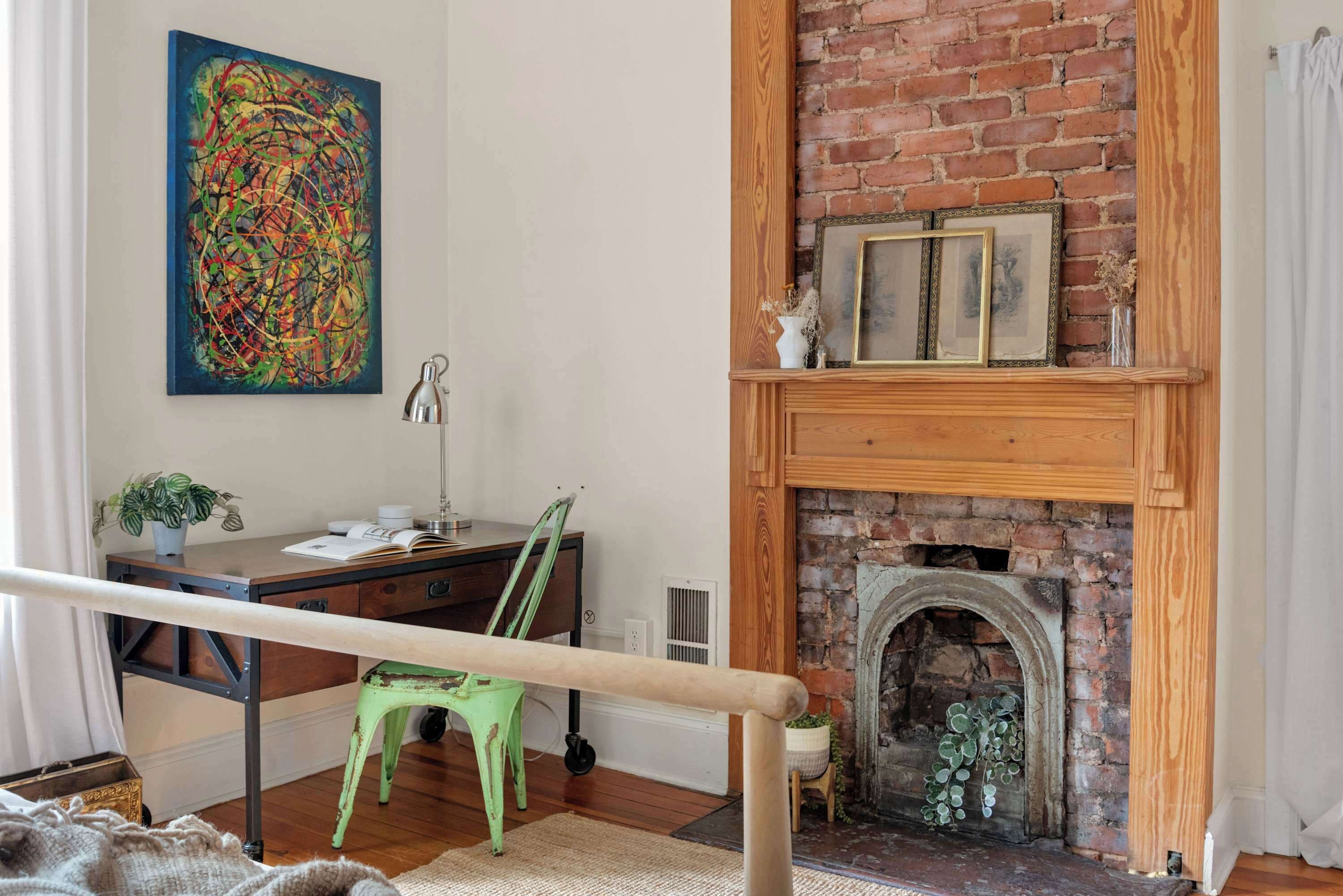 A room with a brick fireplace, a desk with a chair, a green chair, a potted plant, and a colorful abstract painting on the wall.