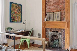 A room with a brick fireplace, a desk with a chair, a green chair, a potted plant, and a colorful abstract painting on the wall.