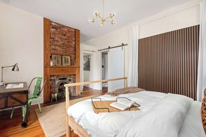 Um quarto com cama de madeira, lareira de tijolo, escrivaninha com cadeira e vista para uma porta.