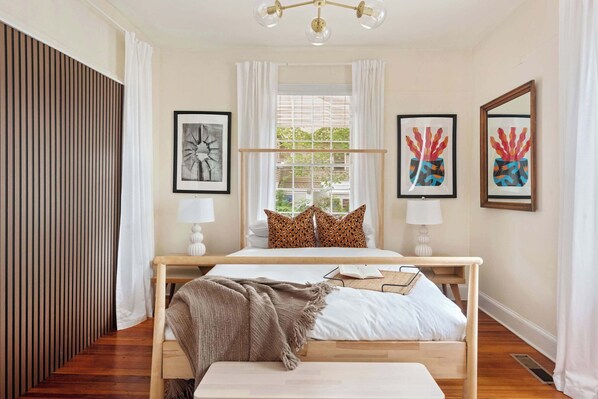 A bedroom with a wooden bed, white bedding, a bedside table with a lamp, and two framed artworks on the wall.