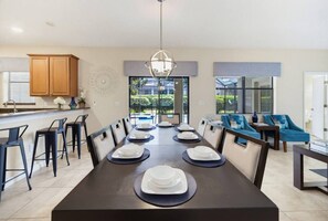 Comedor con una mesa larga dispuesta para seis personas, rodeada de sillas, con cocina y zona de estar visibles al fondo.
