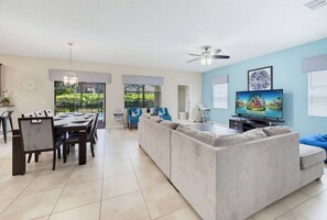 A modern living room with a dining area, a large sofa, a flat-screen TV, and a ceiling fan. - 6BR Family Friendly Resort Pool Home Near Disney (Davenport)