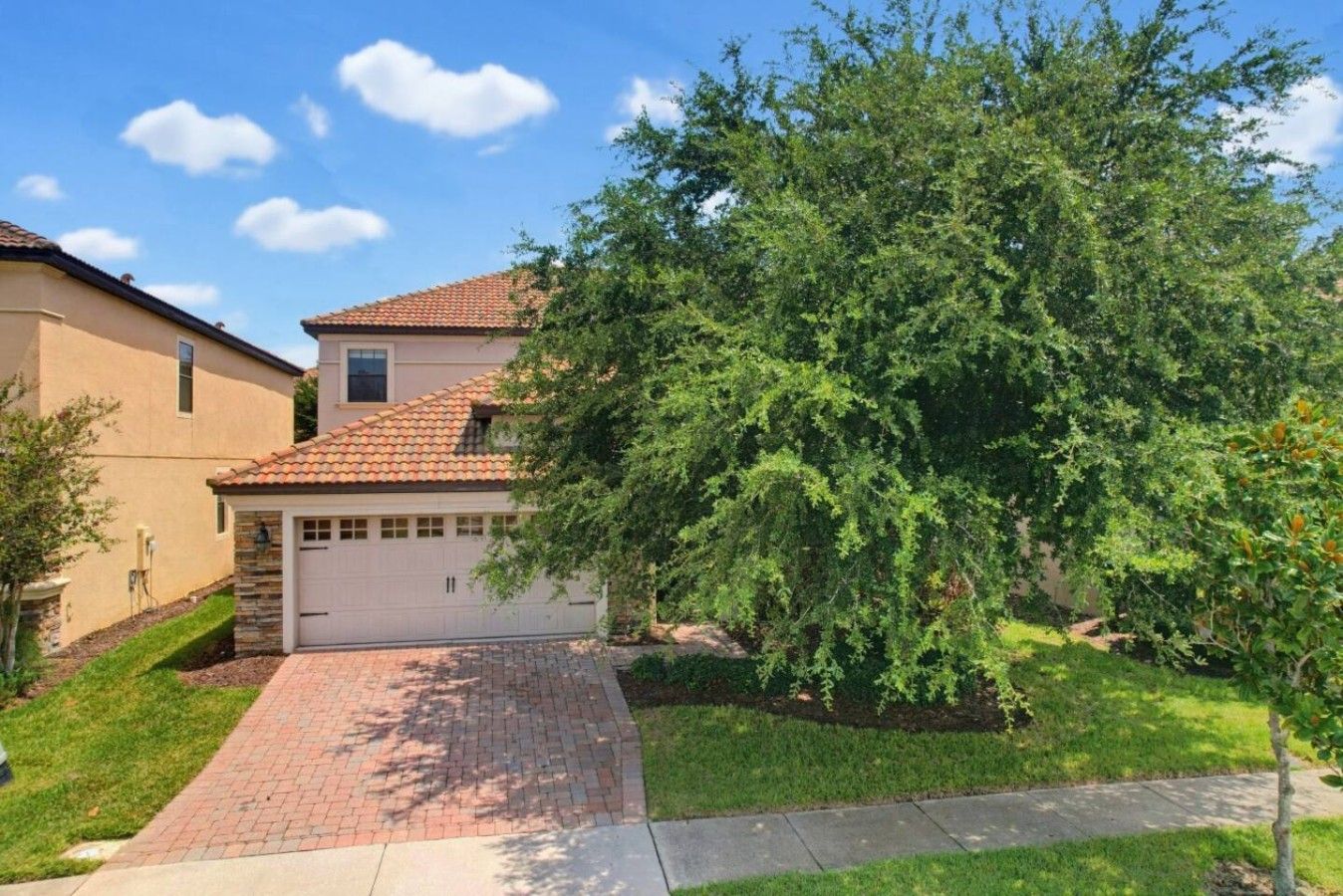 A residential house with a garage, a brick driveway, and a large tree in the front yard.