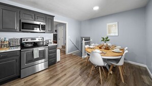 Una cocina moderna con electrodomésticos de acero inoxidable, un comedor con mesa de madera y sillas blancas, y una pared gris con un cuadro enmarcado.