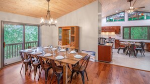 A spacious dining area with a wooden table set for six, surrounded by chairs. A large wooden cabinet is visible in the background, and the room has large windows offering a view of the outdoors. - Private Mountain Home, 2miles To Ski, Hot Tub, Views, Fireplace, & Pet Friendly! (Beech Mountain)