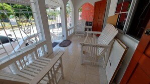 Uma área de estar coberta ao ar livre com cadeiras brancas e uma mesa, uma porta vermelha e vista para a rua.