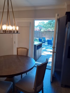 A dining area with a round table and chairs, a kitchen with a refrigerator, and an open door leading to an outdoor patio with chairs and a table.