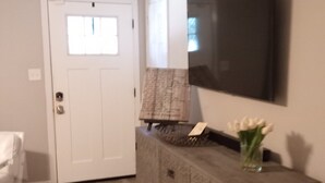 A room with a wooden floor, a white door, a flat-screen TV mounted on the wall, and a small table with a vase of flowers. - Charming Home Comfy Beds Private Backyard with Fire Pit  (Hattiesburg)