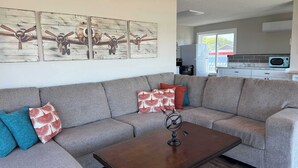Un salón con un sofá seccional gris, cojines estampados rojo y blanco, una mesa de centro de madera y una obra de arte en la pared con aviones antiguos.